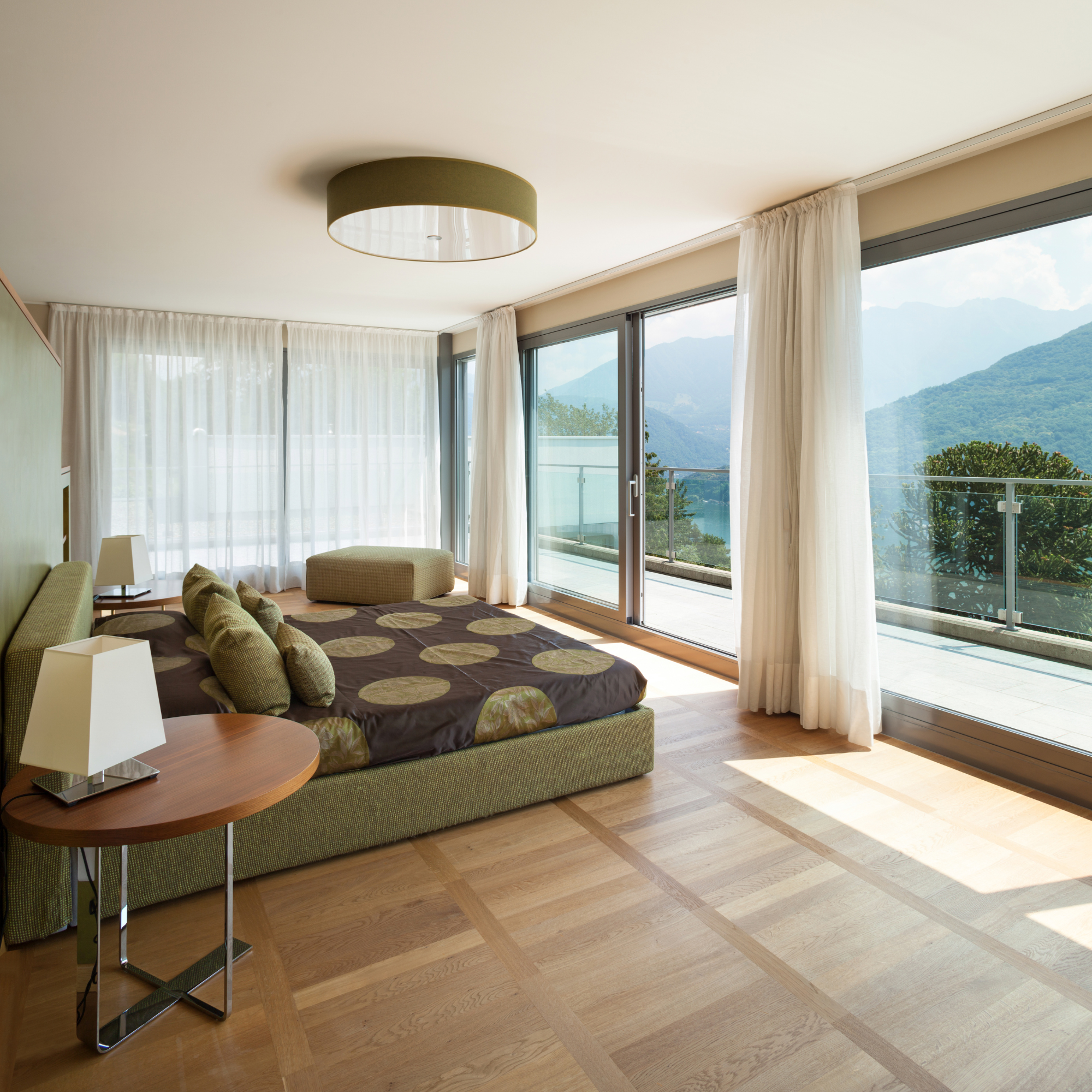 A modern living room with a large sliding glass door opening to a balcony with a mountain view. The room features sheer white curtains, a green upholstered sofa with throw pillows, a round ottoman, a wooden side table, and a ceiling-mounted light fixture.