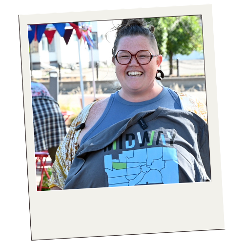 A young woman with glasses and dark hair in a bun, smiling outdoors at a festive event with bunting in the background, carrying a backpack and wearing a blue shirt.