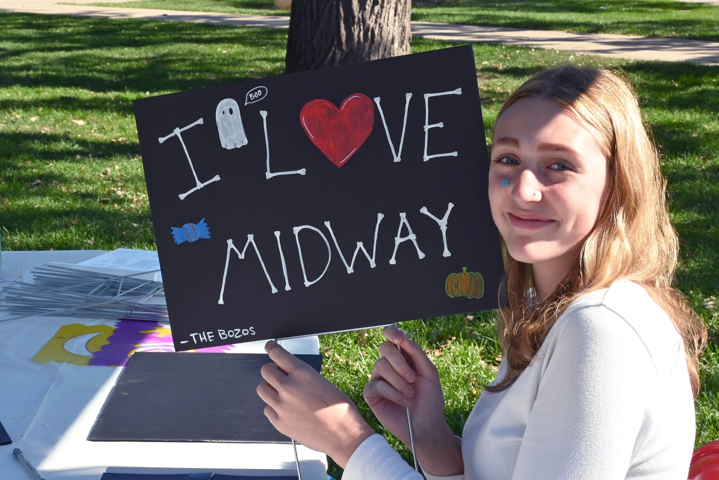 A young girl with long light brown hair holding a black sign with white painted letters spelling "I LOVE MIDWAY" with a red heart in place of the word "love". She has a blue star sticker on her nose and is smiling. The background shows a park with a 