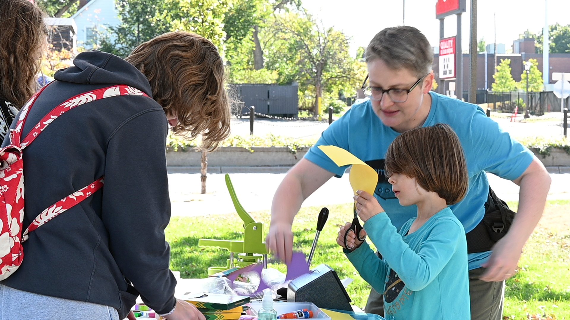 A group of four people, including two children, gathered outdoors around a table with various craft supplies. They are working on a craft project, with trees and buildings in the background, suggesting a park or urban outdoor setting.