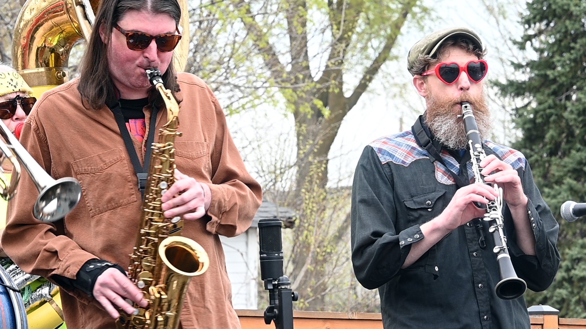 Two men playing saxophone and clarinet outdoors during daytime, with trees and a house in the background. Both men wear sunglasses, one with heart-shaped red sunglasses and a hat, the other with classic sunglasses.
