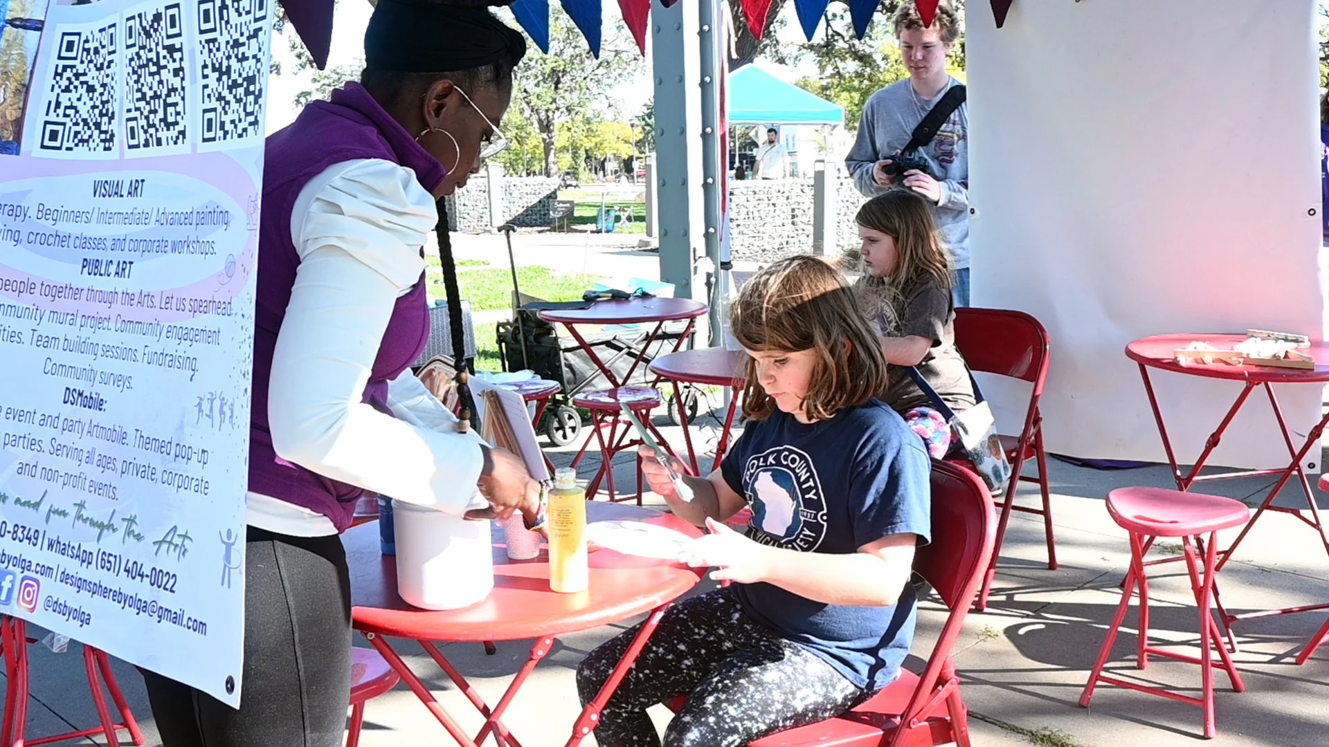 A woman at a table shows children artwork. Four children, three girls and one boy, are engaged with their artwork at an outdoor event. There are red chairs and tables, a large white panel, and a QR code on a sign in the foreground. The background fea
