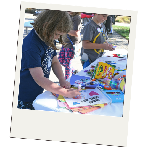 Children at an outdoor table engaging in arts and crafts, with various supplies like markers, paper, and Elmer's glue.