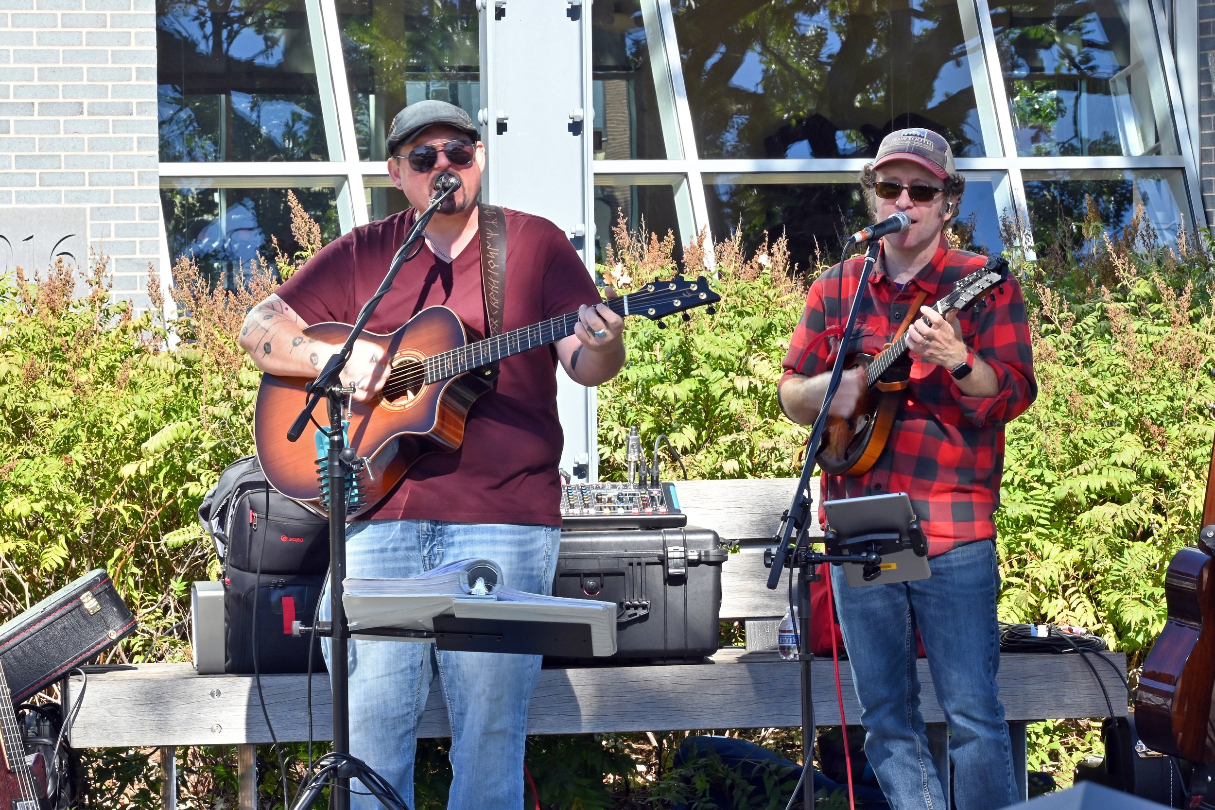 Two men perform as a band outdoors. One man plays an acoustic guitar and sings into a microphone, wearing sunglasses, a cap, and a maroon shirt. The other man plays a mandolin and sings into a microphone, wearing glasses, a baseball cap, and a red an