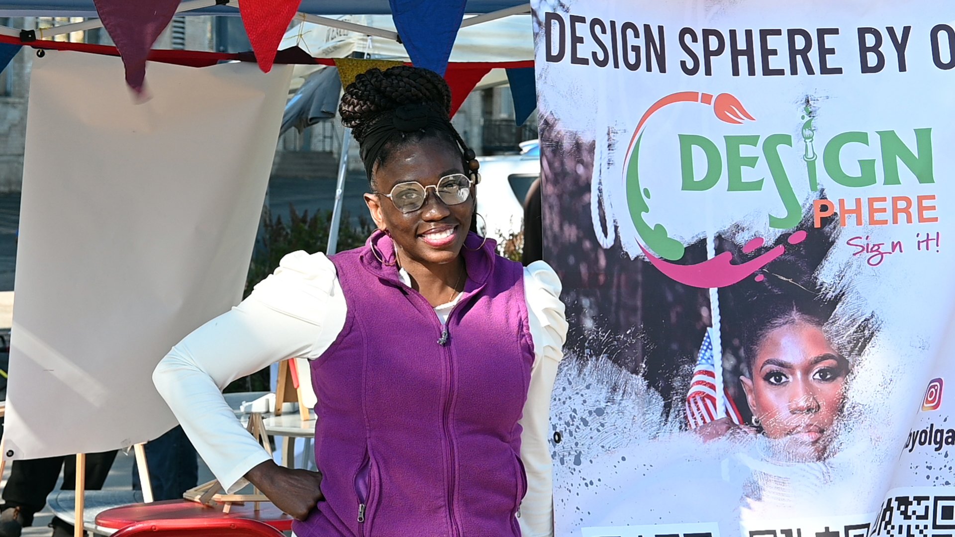 A woman with glasses and braided hair standing outdoors next to a colorful banner that reads 'Design Sphere' with artistic graphics and images of other women.