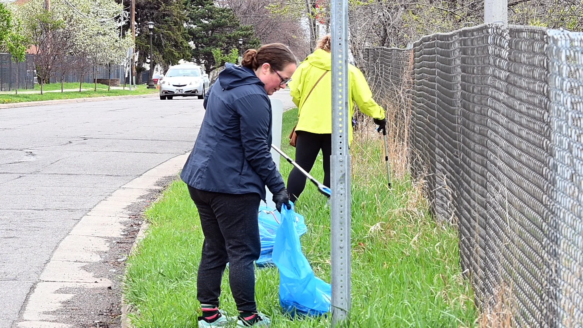 Two women collect trash along a grassy sidewalk during daytime, one wearing a blue jacket and the other a yellow jacket, with black gloves, near a chain-link fence.