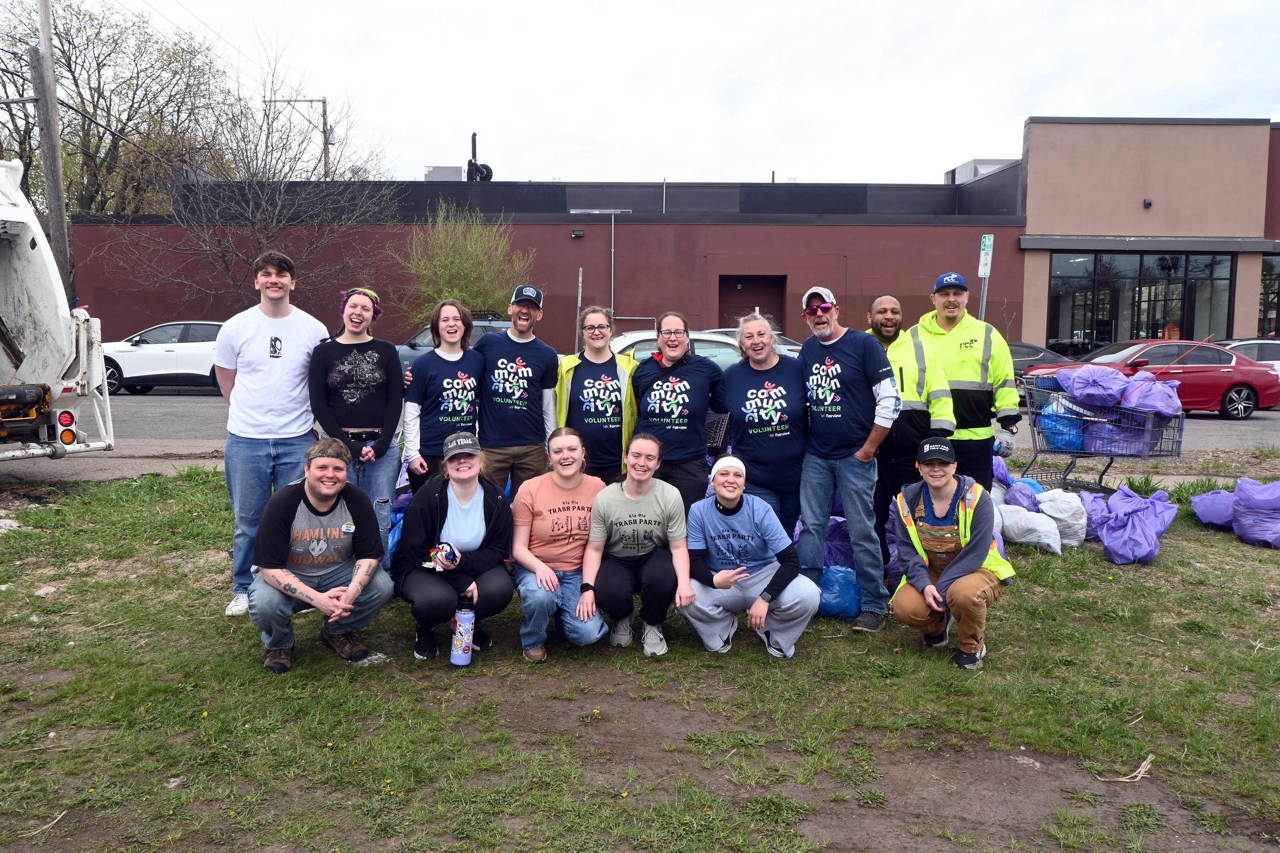 A group of smiling volunteers, both men and women, posing outdoors in front of a building after participating in a community cleanup event. They are surrounded by piles of filled trash bags and a shopping cart filled with more trash. Some are wearing