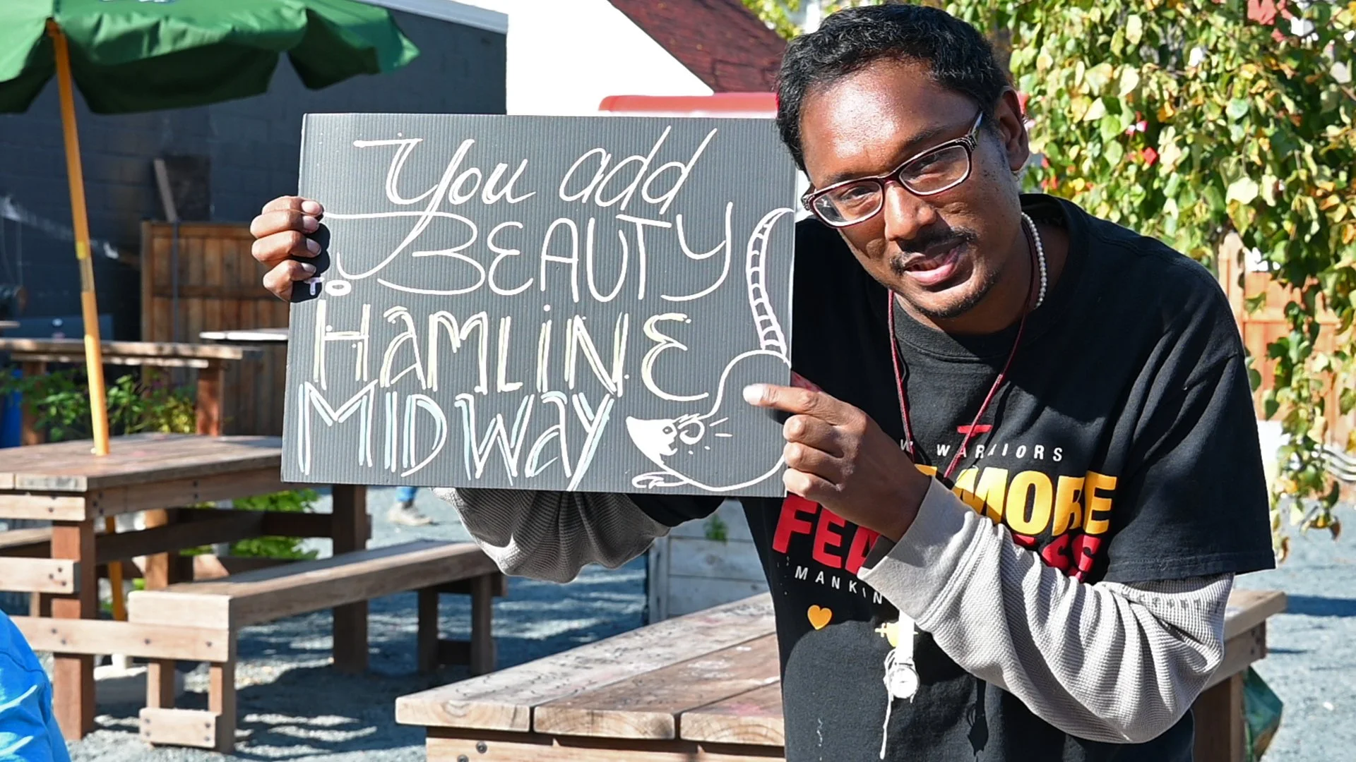 A man with glasses and a black hoodie is holding a sign that says 'You add beauty to Hamilne Miday.' He is standing outdoors with trees and wooden benches in the background.