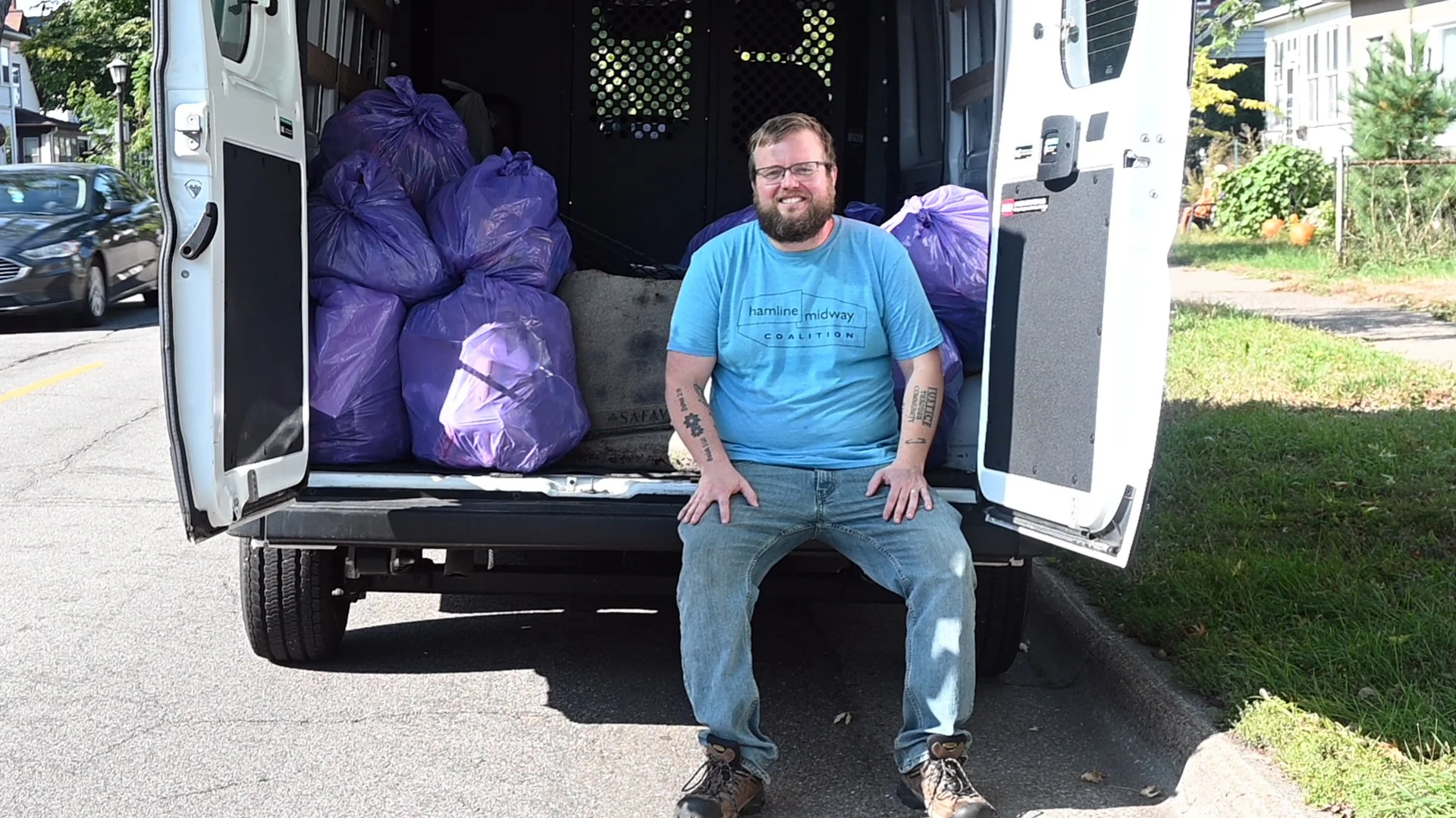 A man with glasses and a beard sitting on the edge of an open van filled with large purple garbage bags, smiling at the camera.