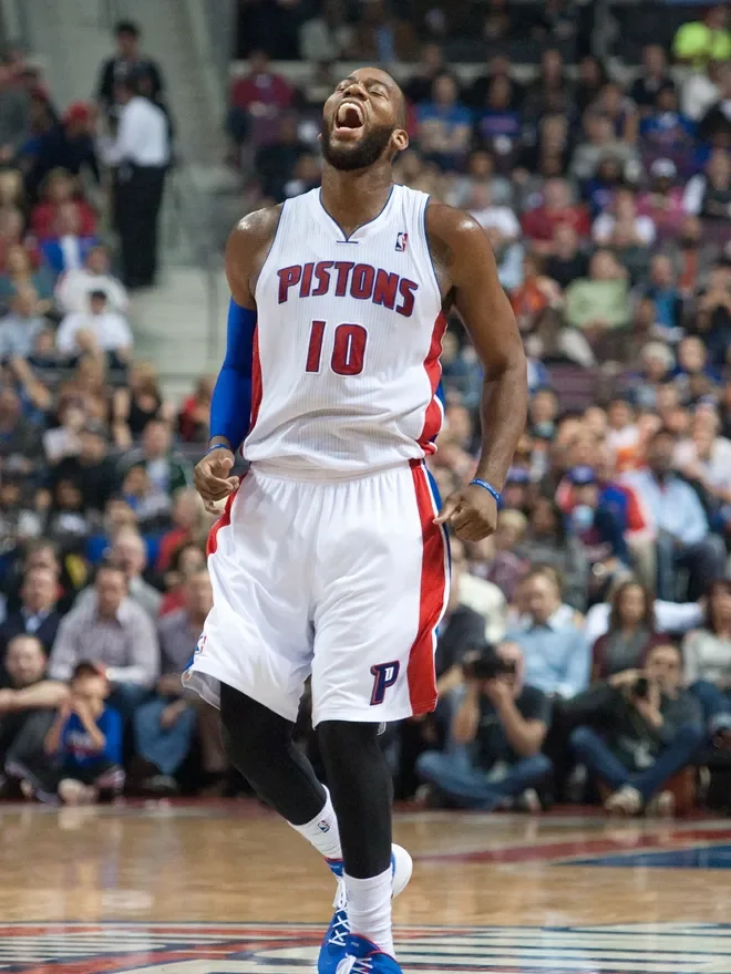 A basketball player wearing a Detroit Pistons jersey with number 10 celebrating on the court, with an audience in the background.