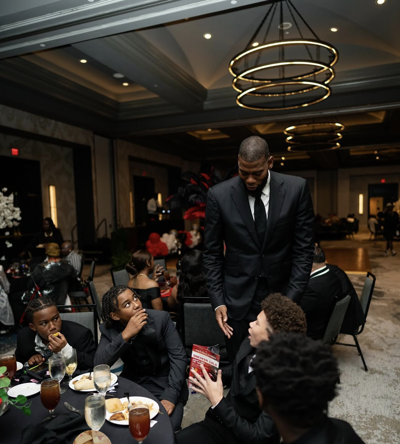 At a formal event, a man in a black suit is standing and talking to a seated woman holding a card, with children and other guests around them in a decorated banquet hall.