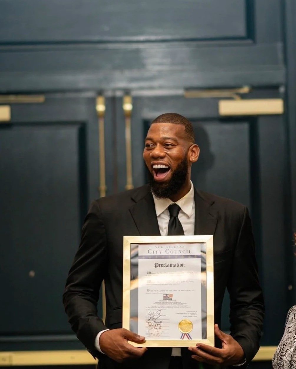 A smiling man in a black suit holding a framed proclamation certificate from New Orleans City Council, standing in front of dark blue doors.