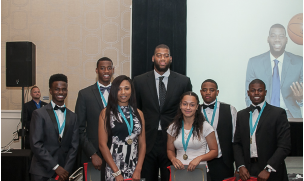 Group of young adults dressed in formal attire with medals around their necks, standing on a stage after an award ceremony, with a large screen behind them displaying a photograph of an older man in a suit.