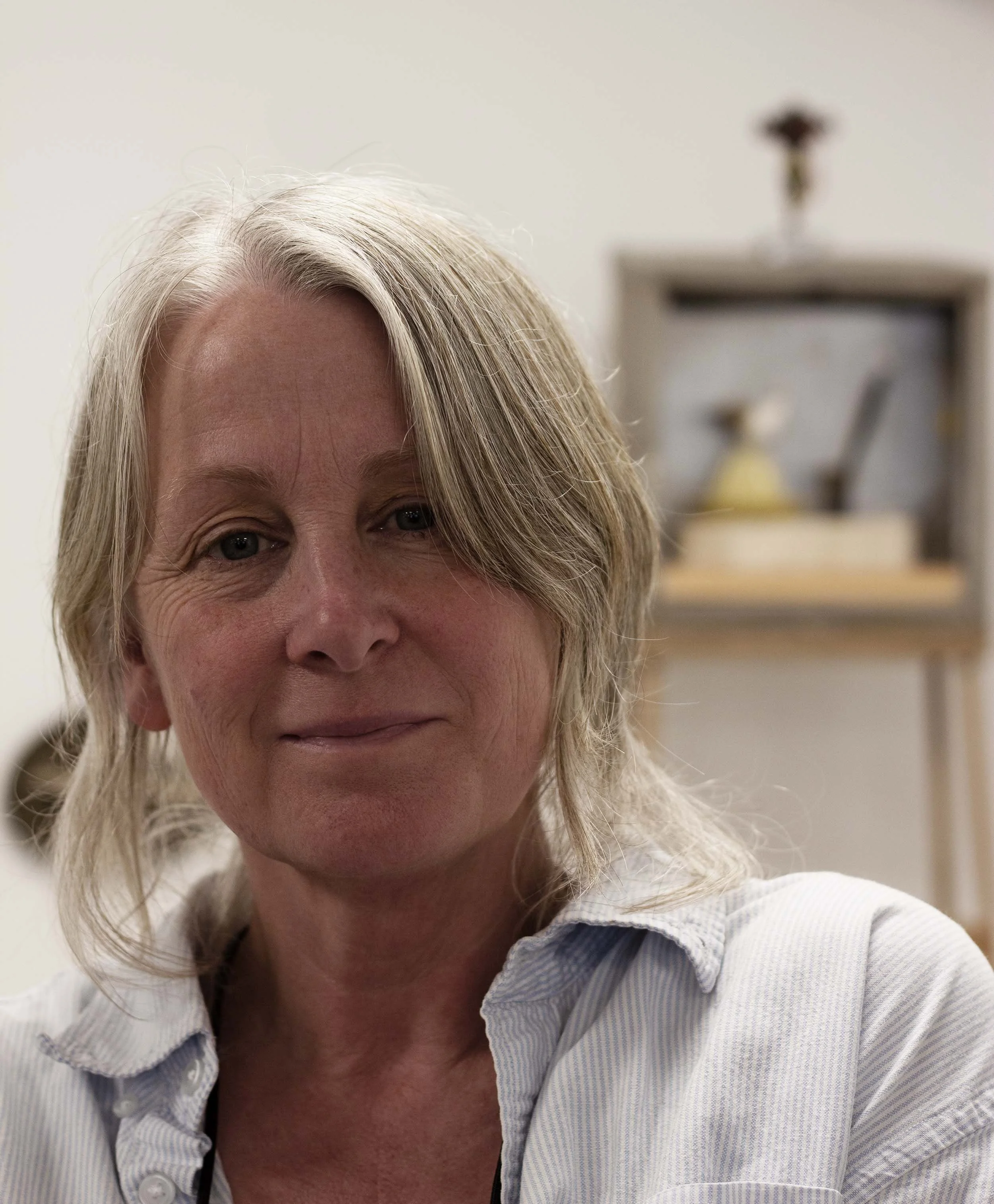 Close-up of the artist Stephanie Chaldecott, wearing a white collared shirt with thin blue stripes, smiling gently, with a blurred background of her studio..