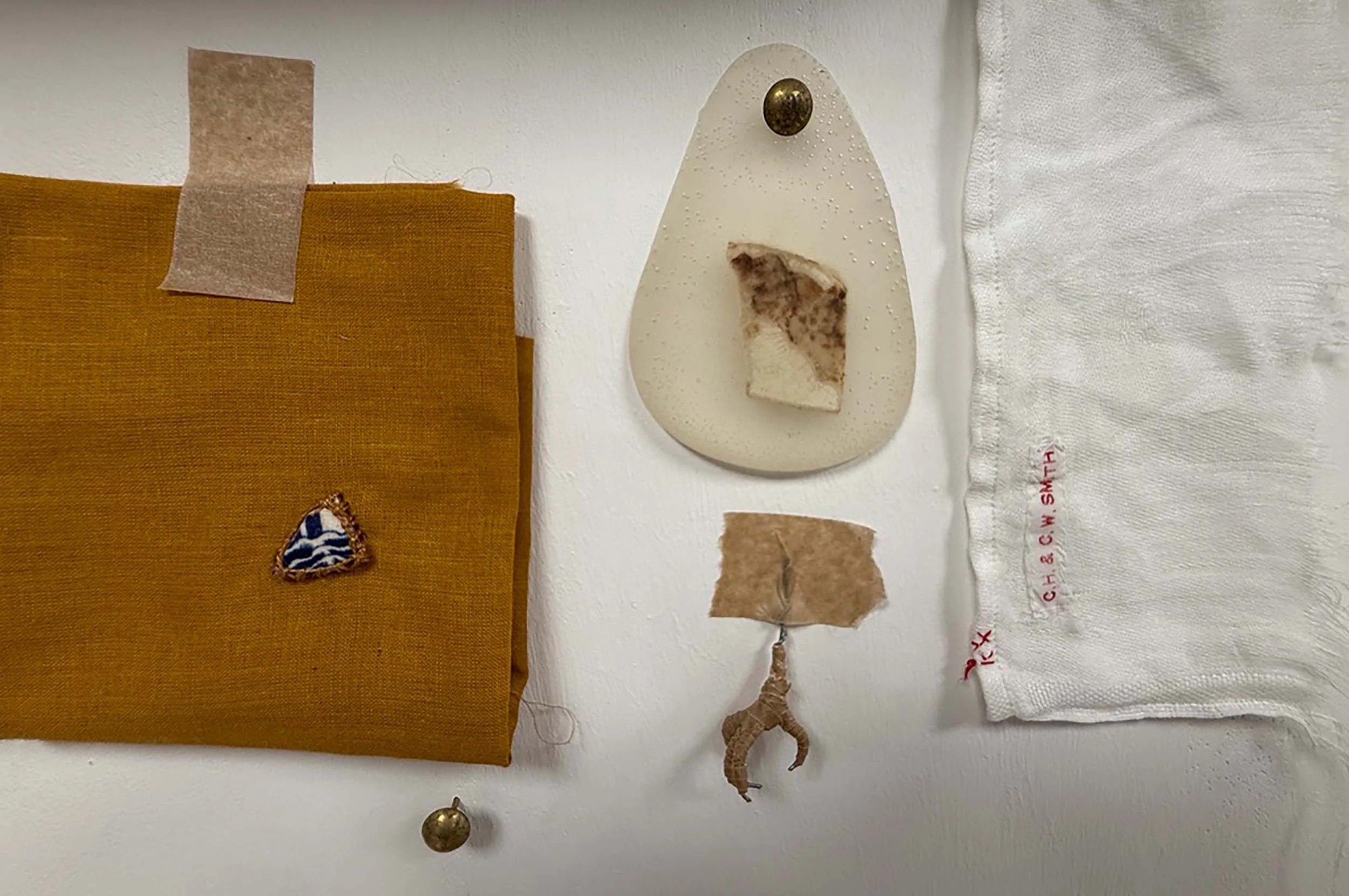 The image shows a white wall with various objects attached, including a mustard yellow fabric piece with a decorative butterfly patch, pieces of torn brown paper, a white cloth with red embroidery, a teardrop-shaped object with a dark spot, a small round metallic object, and a piece of regulation document.