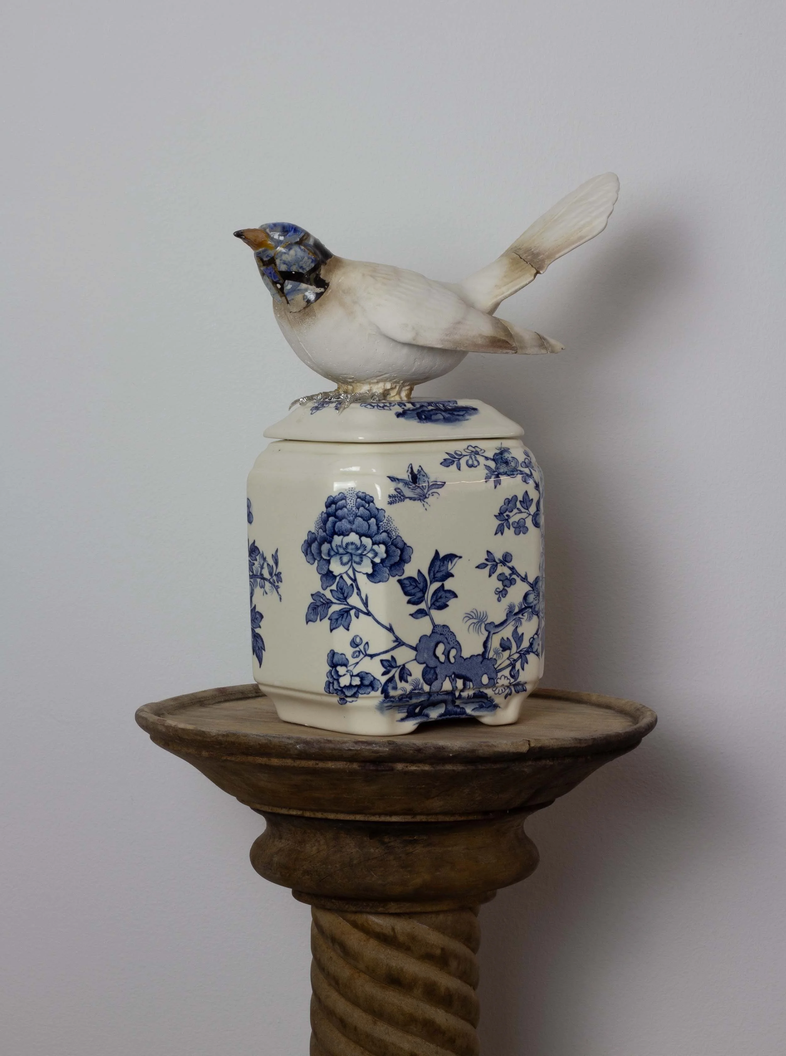 A decorative sculpture of a bird, made from ceramic or similar material, perched on a blue and white patterned vase that is placed on a wooden stand.