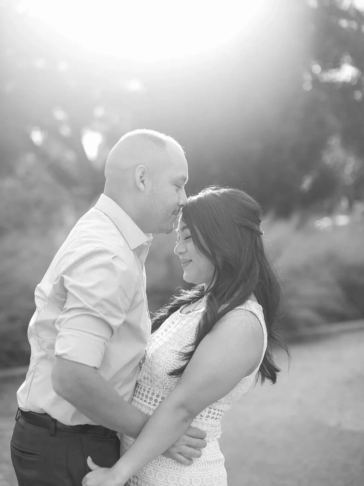 A black and white photo of a couple in love, standing close with foreheads touching, in an outdoor setting during golden hour, with trees blurred in the background.