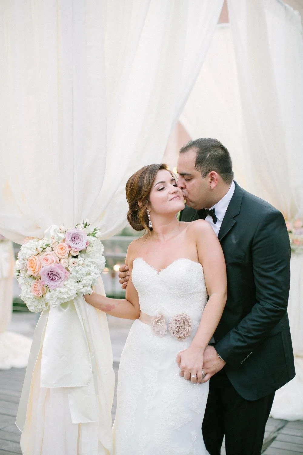 A bride and groom share a kiss at their wedding, with the bride holding a bouquet of pink, purple, and white roses, and the groom dressed in a black tuxedo.