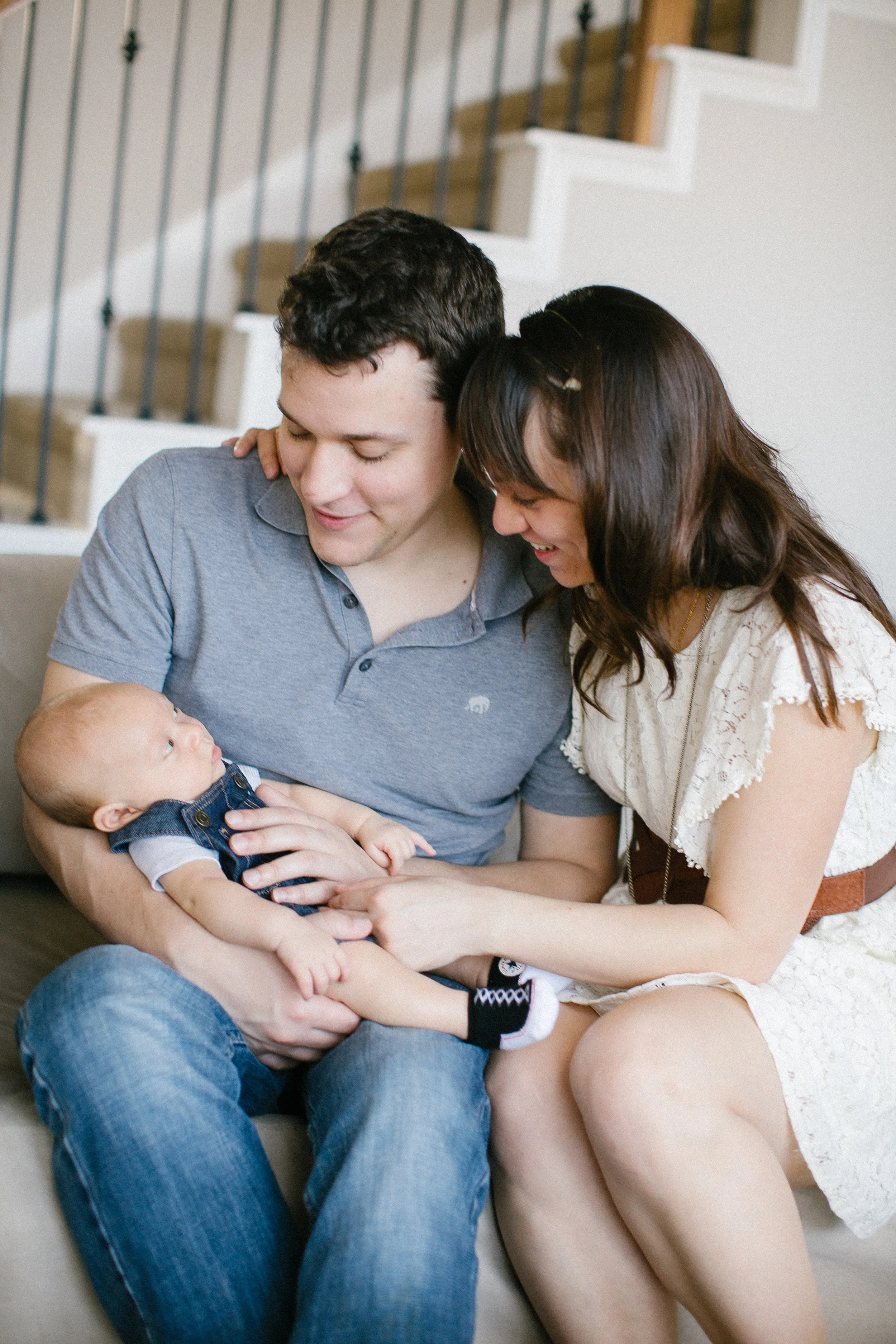 A family sitting on a beige couch in a living room, with a man holding a baby and a woman smiling at the baby.