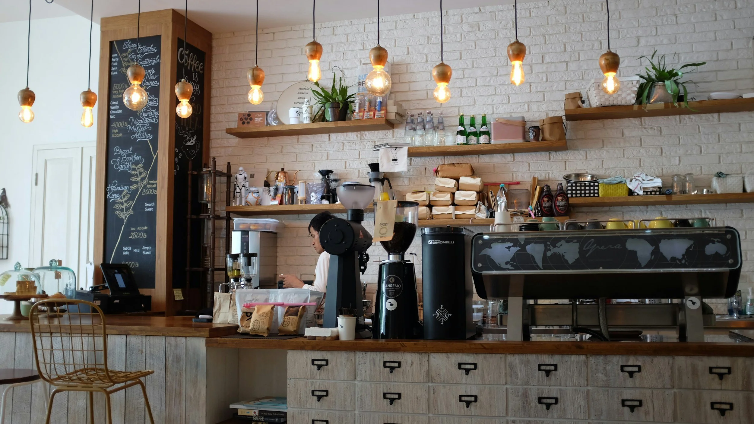 Interior of a coffee shop with hanging Edison-style light bulbs, a barista preparing coffee, shelves with supplies, a menu board, and a world map on the front of the counter.