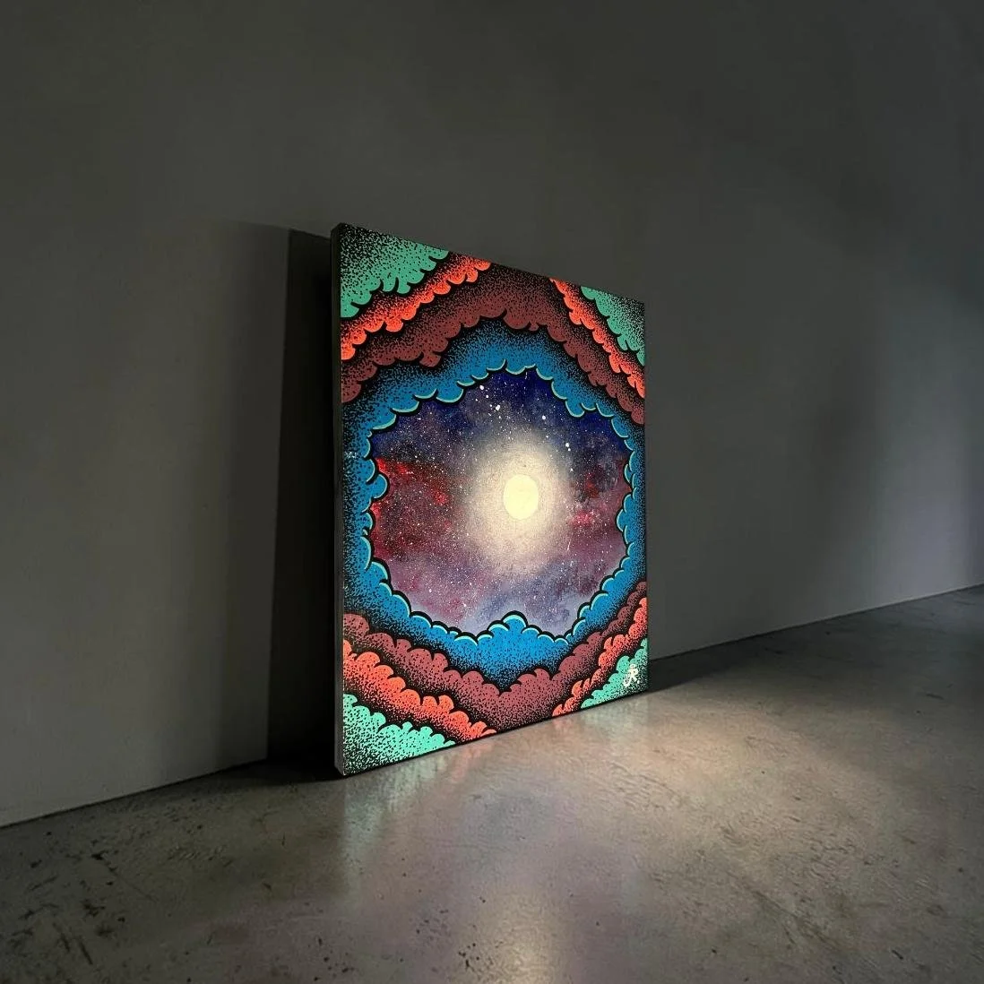 An illuminated painting featuring a cosmic scene with stars, nebulae, and a bright moon or sun in the center, surrounded by colorful, patterned clouds in red, blue, green, and black, leaning against a blank gray wall in a dimly lit space.