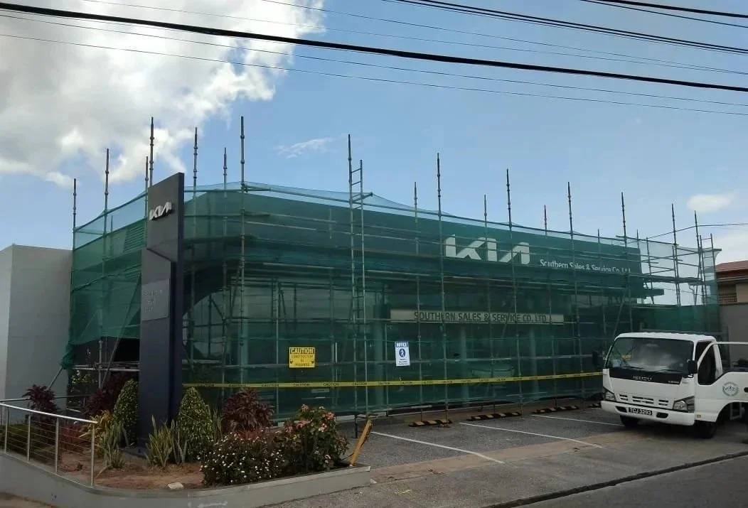 Building covered in green construction netting with scaffolding, a sign for Kia, and a white truck parked nearby, in a parking lot with plants in the foreground.
