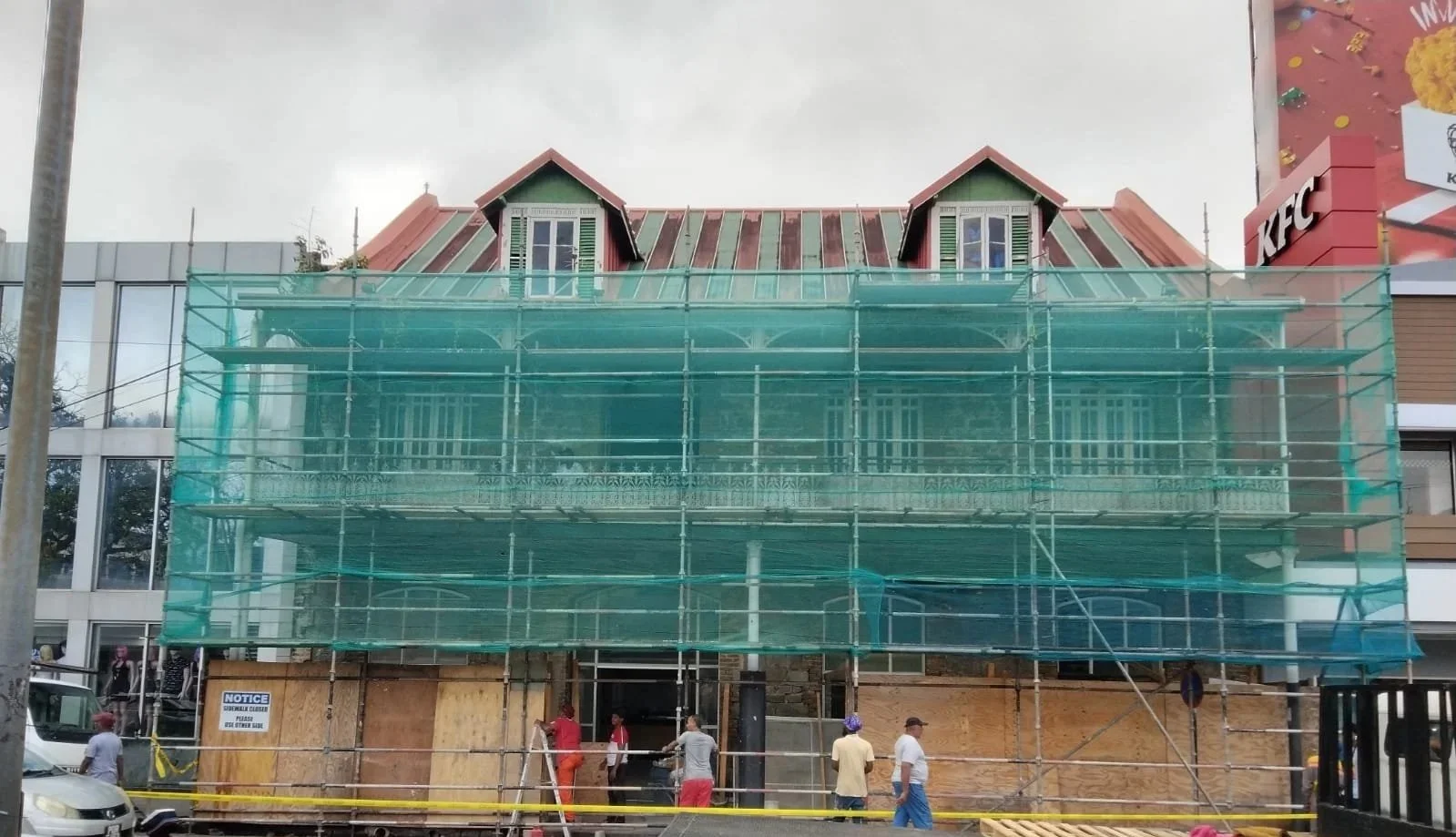 Building under construction with green scaffolding and a wooden boarded entrance, nearby a KFC restaurant.