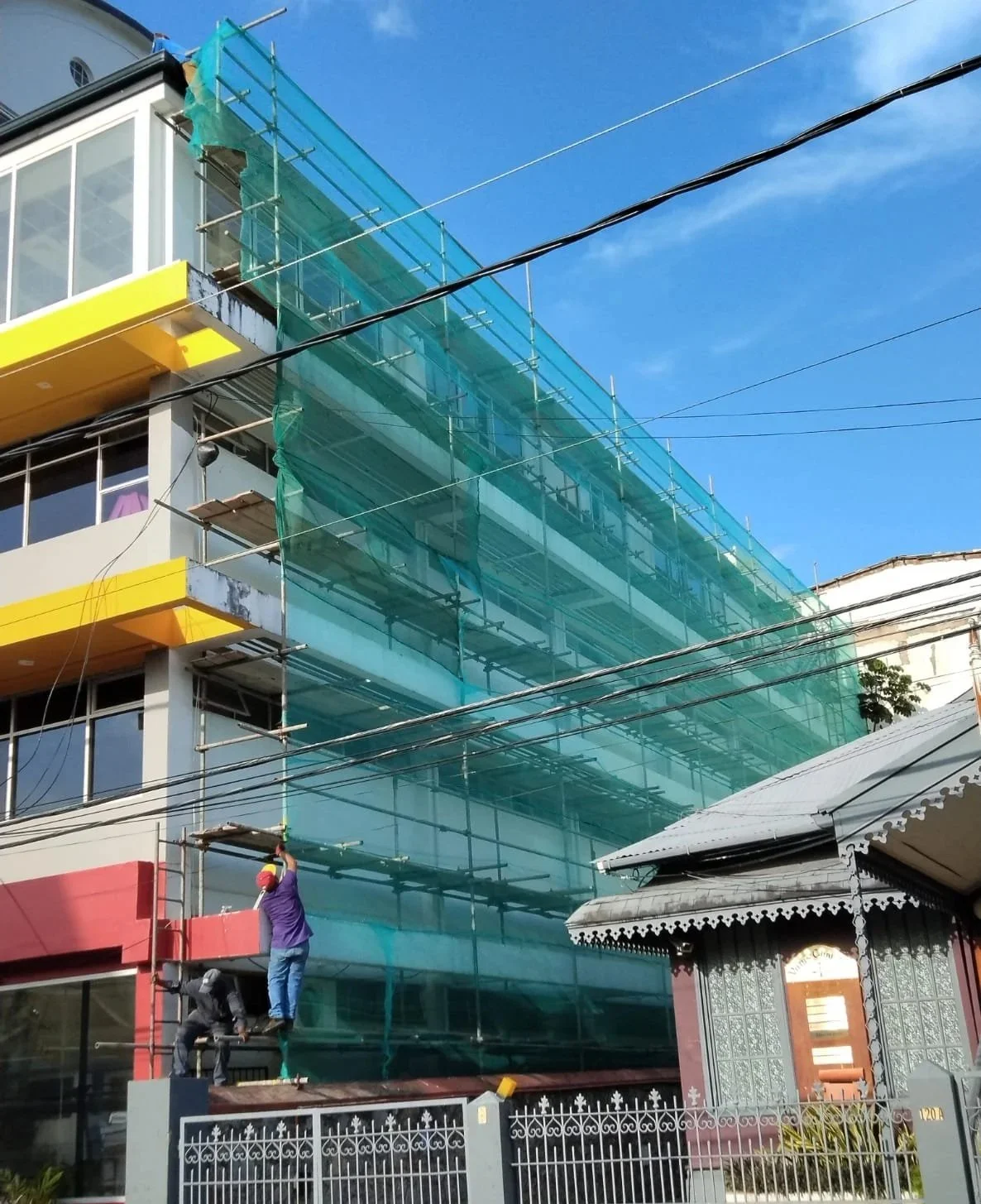 Construction workers working on scaffolding outside a modern multi-story building with glass windows, with a blue sky and clouds in the background.