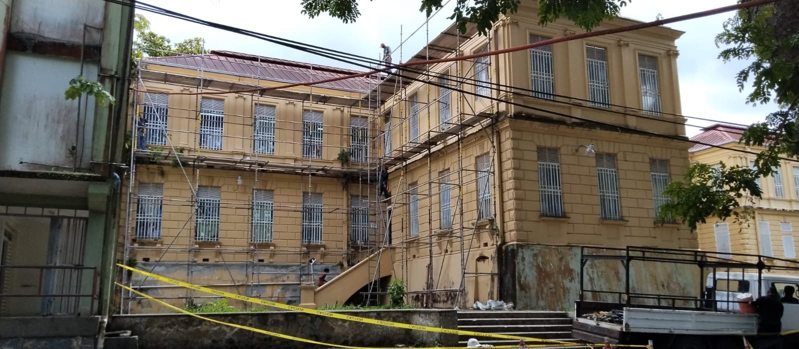 A building undergoing renovation with scaffolding and construction workers on site, and caution tape around the area.