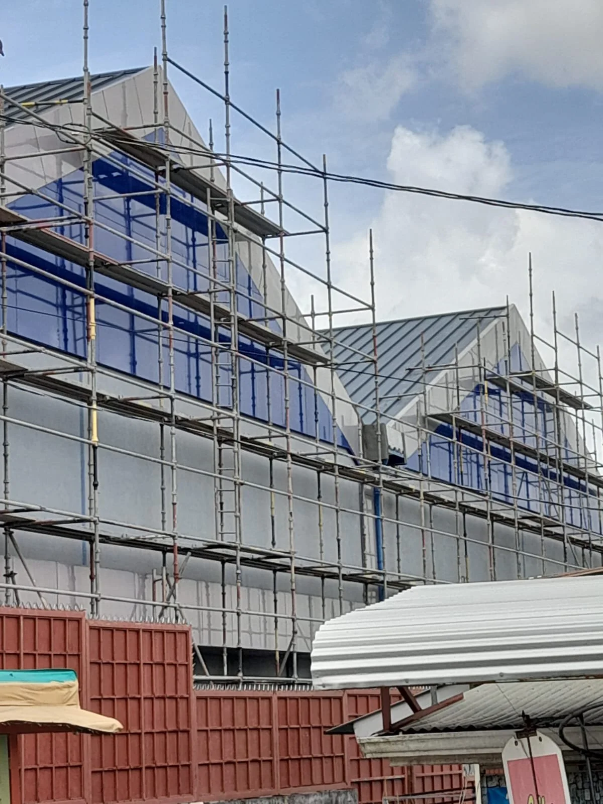 Construction site with scaffolding surrounding a building with blue glass windows and a metal roof, under a partly cloudy sky.