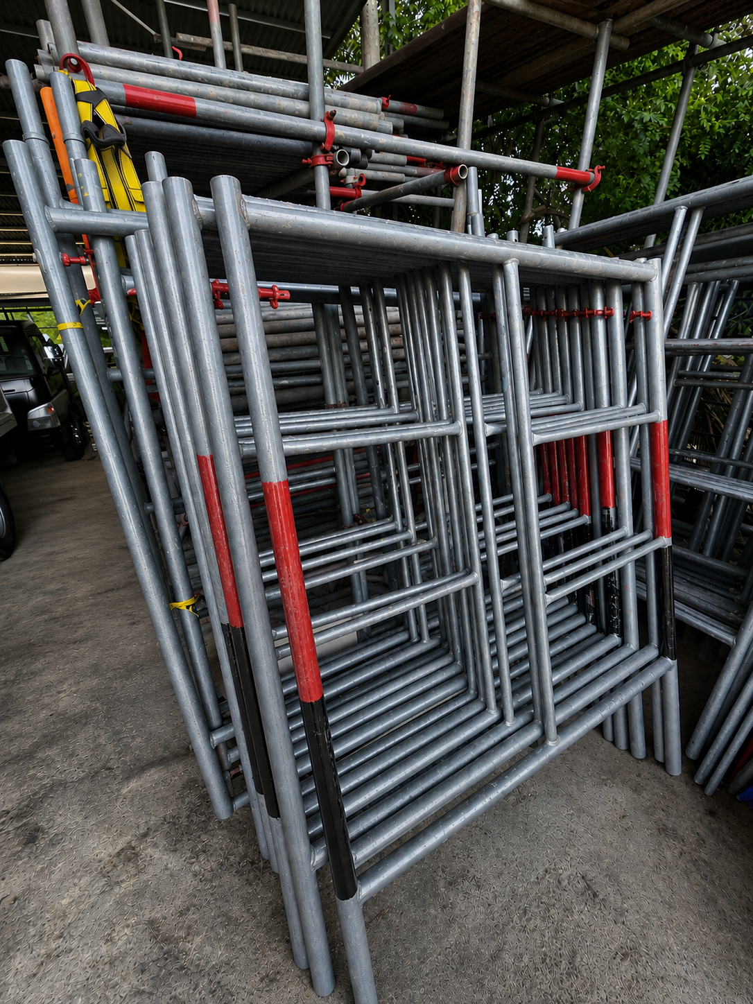 Stacked metal scaffolding frames stored outdoors, with some painted red and black accents, in a construction or hardware supply yard.