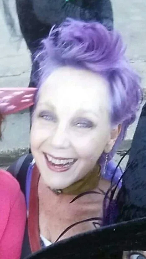 A woman with short, wavy purple hair smiling and looking at the camera.