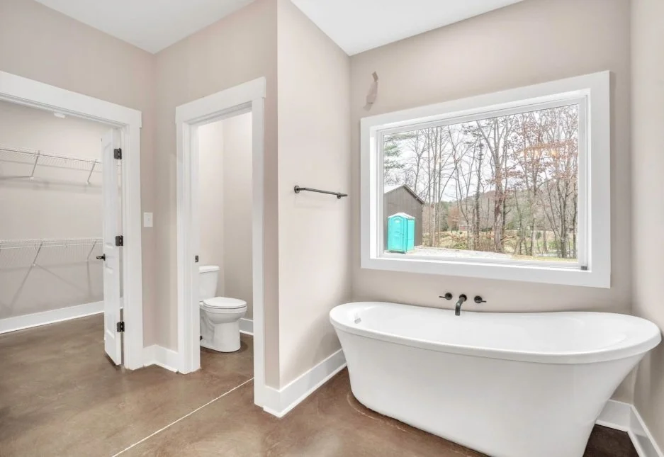 Bathroom with a white freestanding bathtub under a large window overlooking a yard with trees, an open closet with wire shelving, and a separate toilet area in a small room.