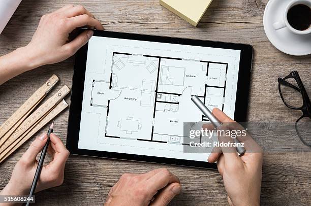 Digital floor plan of a house on a tablet being reviewed by two hands with a stylus and pen, on a wooden desk with rulers, glasses, a cup of coffee, and yellow sticky notes.