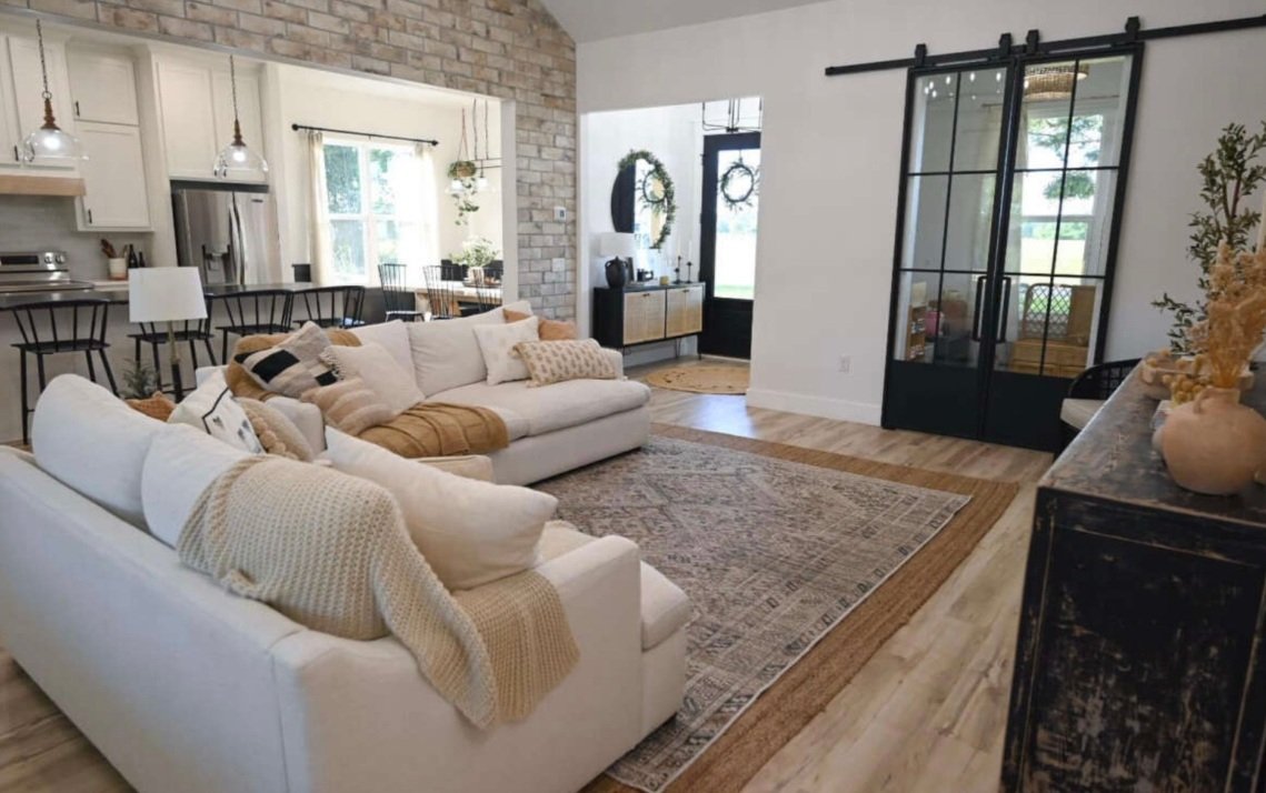 Open-concept living room with white sofas, a patterned area rug, black sliding barn doors, and a view into the kitchen and dining area decorated with neutral tones and greenery.