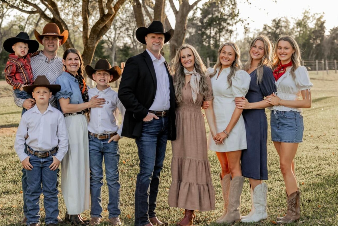 A family dressed in Western attire, including cowboy hats and boots, standing outdoors in a grassy area with trees and a fence in the background.