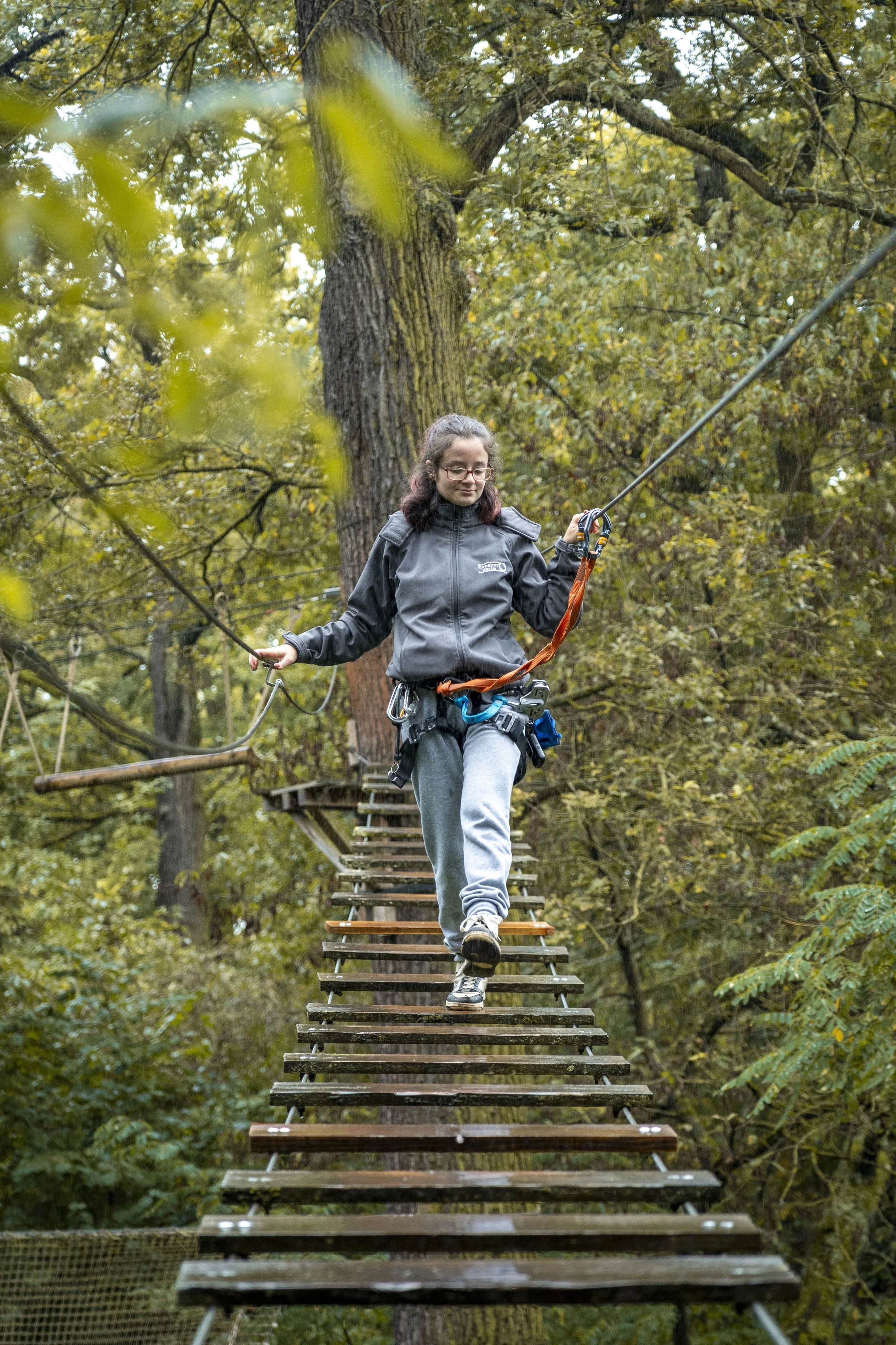 Jeune femme avec cheveux bouclés, lunettes, veste grise, marche sur un pont en bois suspendu dans une forêt dense en automne.