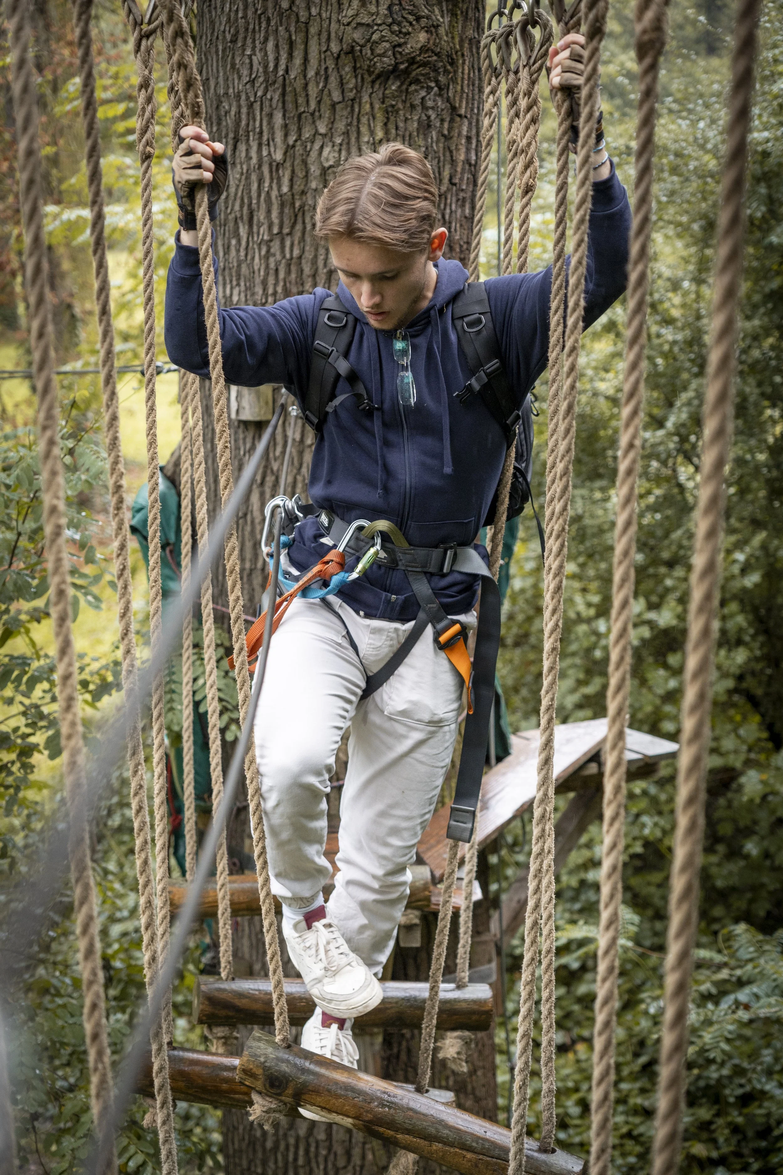 Jeune homme en randonnée, portant un sac à dos, traverse un pont suspendu en forêt, équipé de baudriers de sécurité.