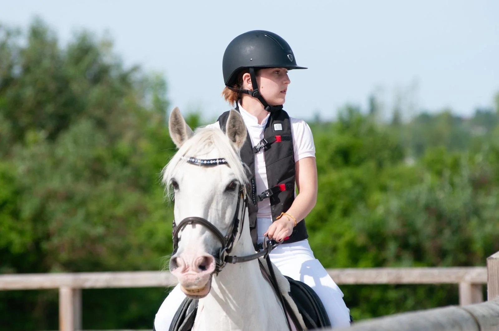 Une jeune femme à cheval, portant un casque noir, une veste noire et un pantalon blanc, en pleine activité équestre dans un environnement verdoyant.