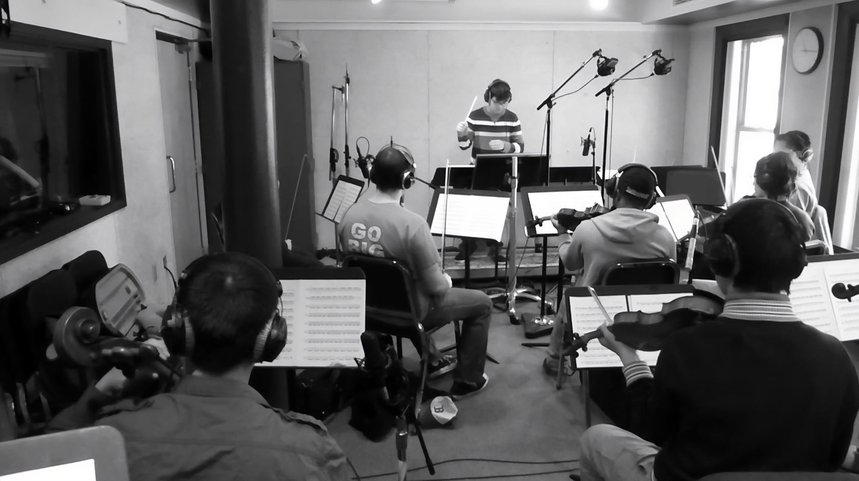 A group of people in a recording studio with music stands and headphones, facing a conductor, Matthew Pablo, standing in front of microphones and equipment.