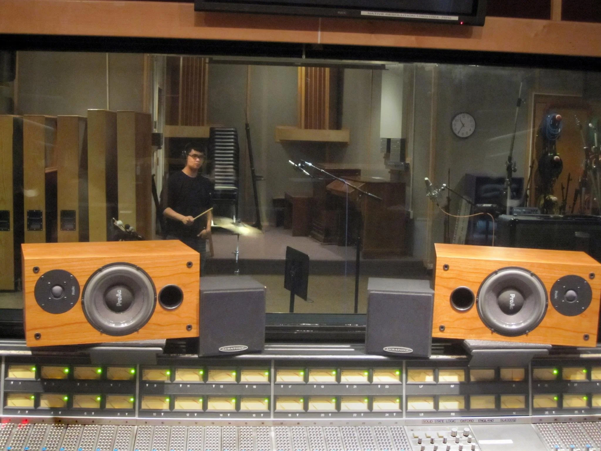 Matthew Pablo holding drumstick in recording studio with audio equipment, large speakers, and soundproofing panels.