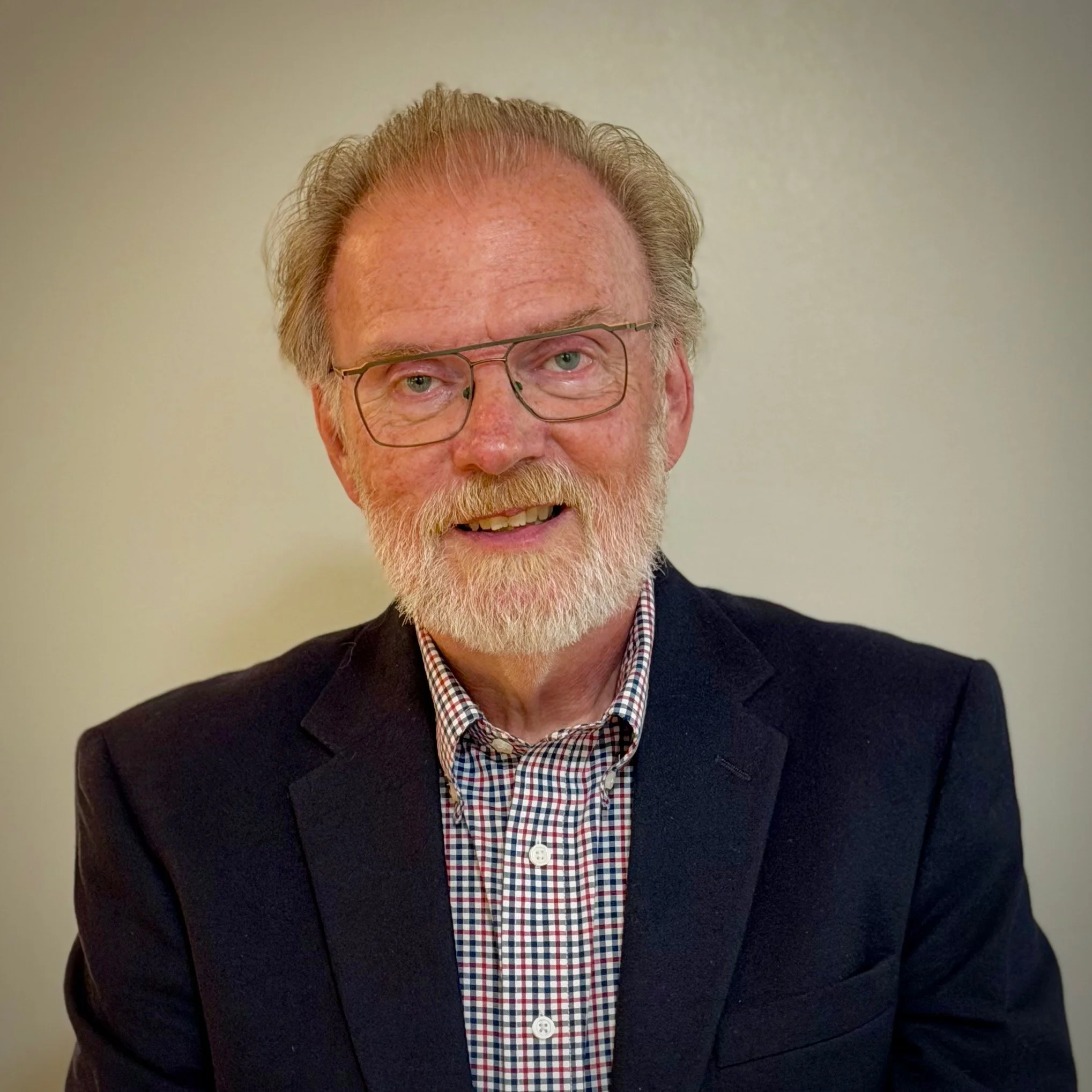 Close-up of a smiling elderly man with gray hair, beard, glasses, wearing a checkered shirt and a dark blazer, against a plain background.