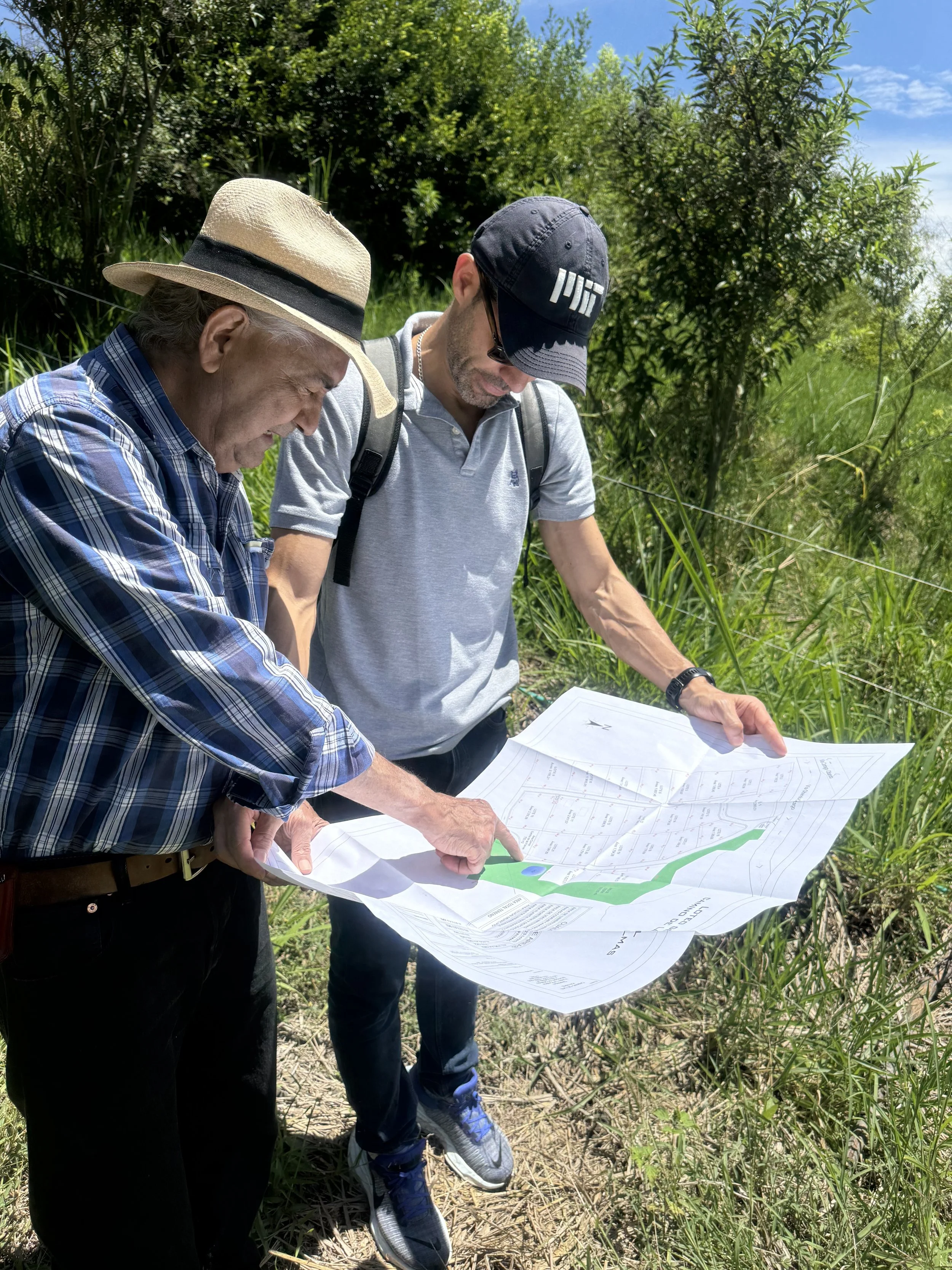 Dos hombres revisando un plano en un entorno natural con vegetación y cielo despejado.