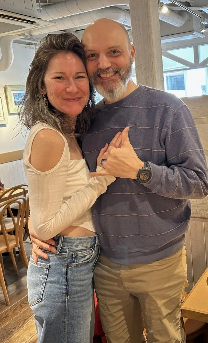 A woman and a man smiling and posing closely together inside a restaurant or cafe. The woman has shoulder-length brown hair and is wearing a cream-colored top with cut-out shoulders, and jeans. The man has a full beard, is wearing a gray striped sweatshirt, khaki pants, and a black watch. They appear happy and friendly.