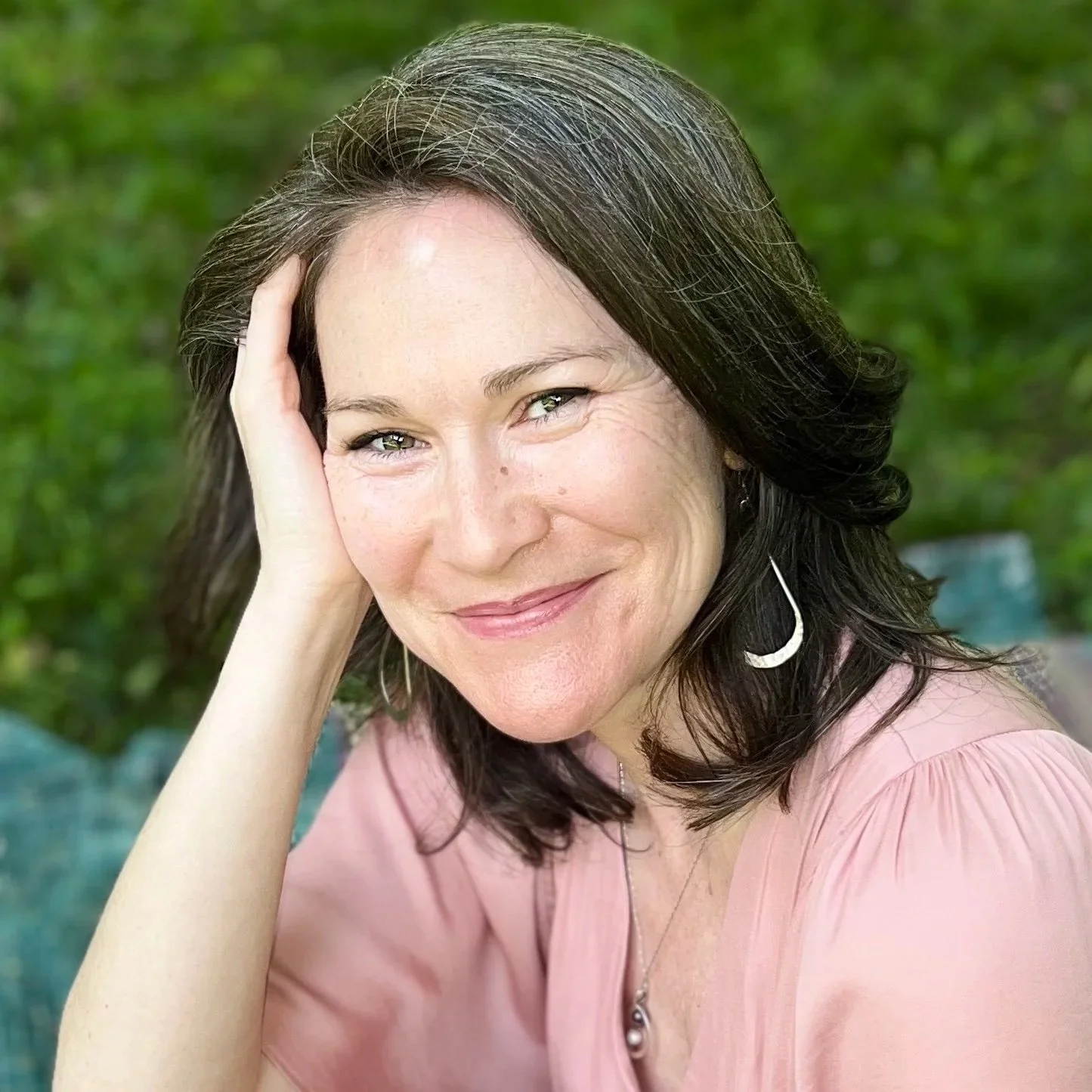 A woman with dark brown hair styled in loose waves, wearing a pink top, silver crescent moon earrings, and a pendant necklace. She is smiling gently, resting her head on her hand, outdoors with green foliage in the background.