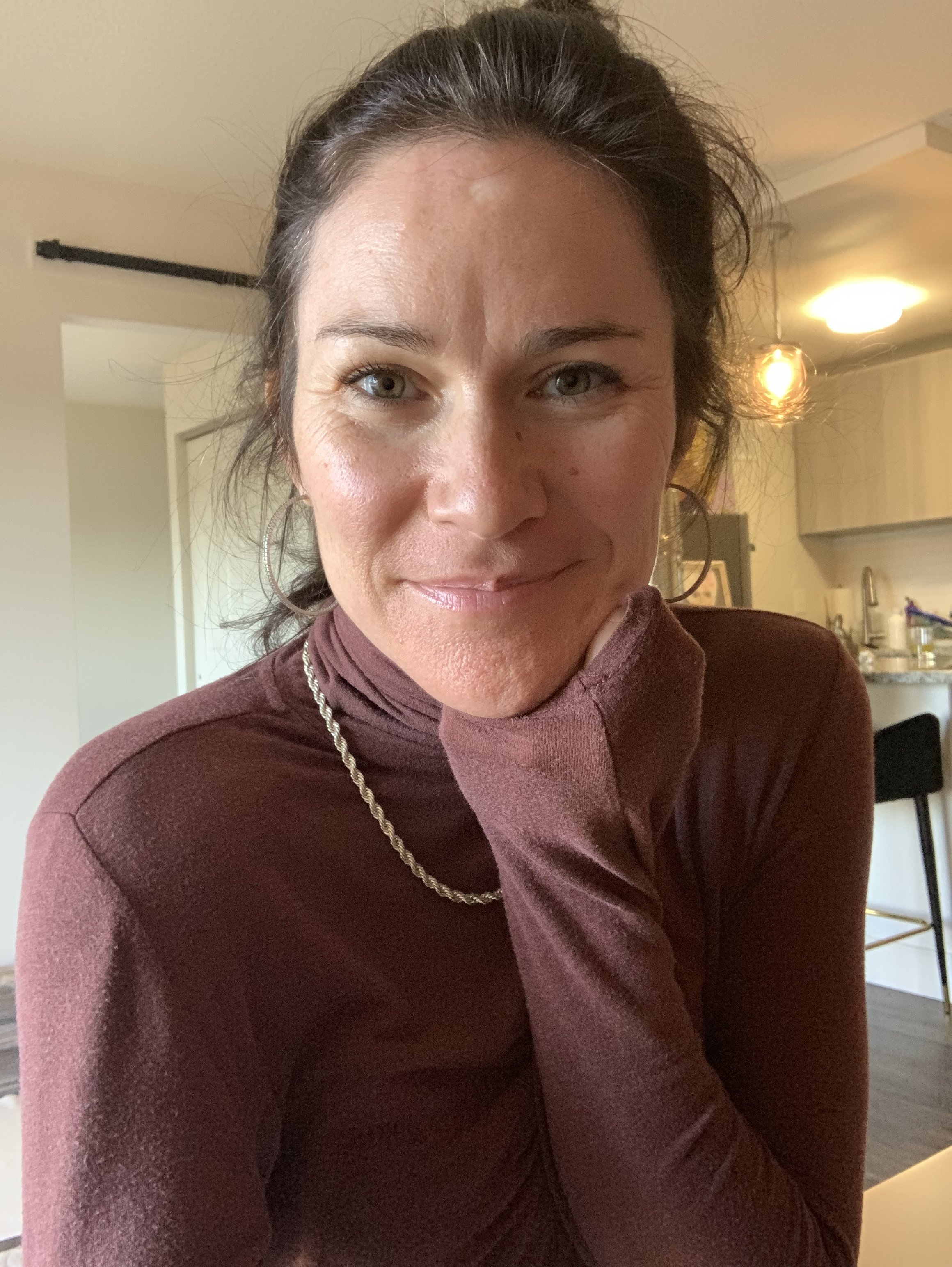 Close-up of a woman with dark hair, wearing hoop earrings and a maroon turtleneck, smiling, in a home kitchen.