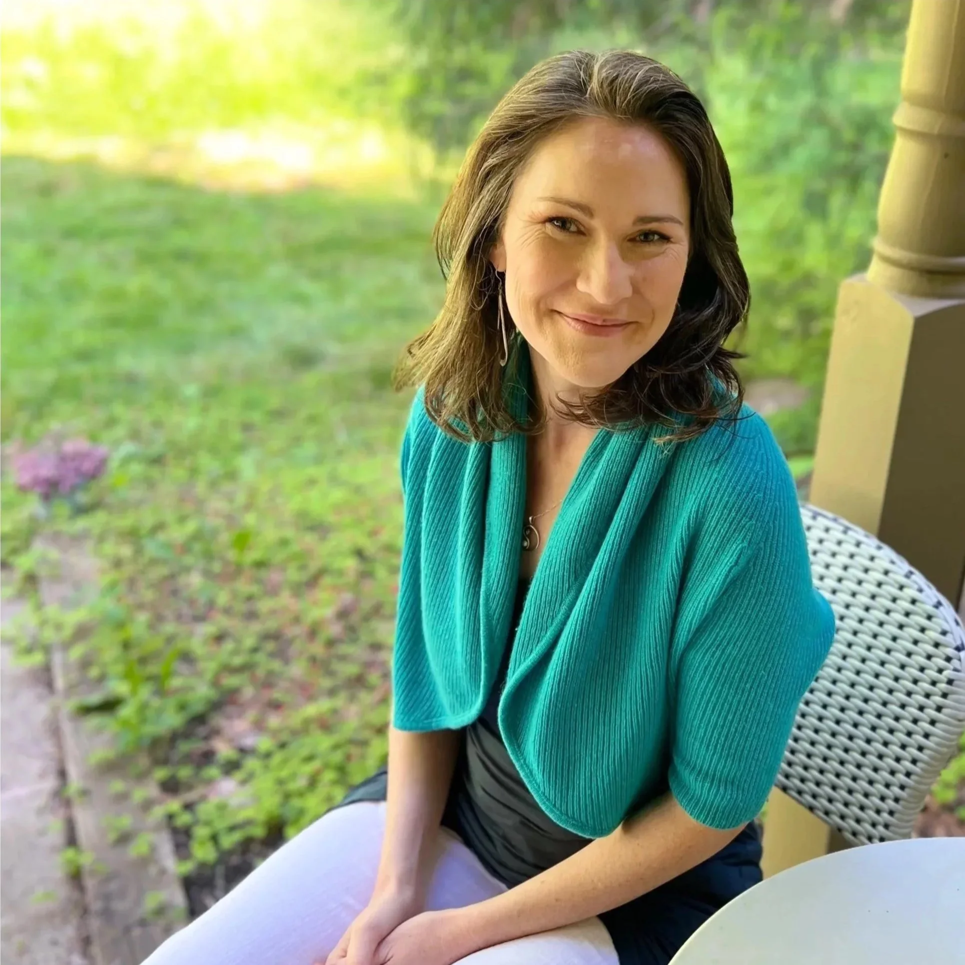 A woman with dark hair and a blue top, smiling outdoors in a green garden, sitting on a wicker chair.