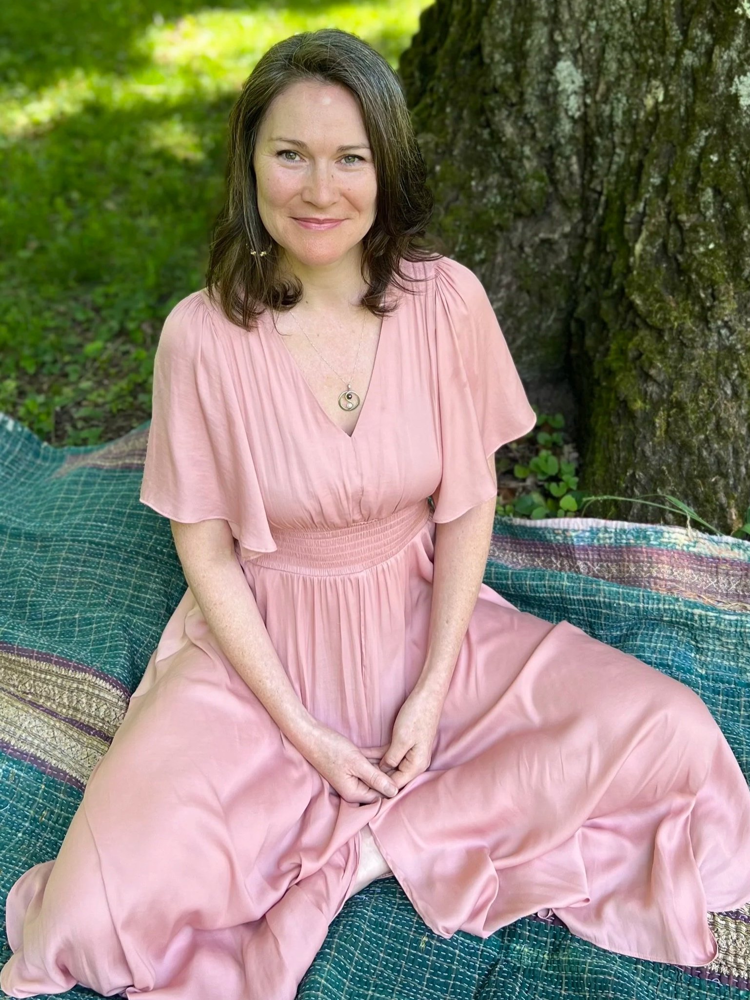 A woman with brown hair and green eyes, wearing a pink dress with a yin-yang necklace, sitting on a colorful blanket outdoors near a large tree, smiling at the camera.