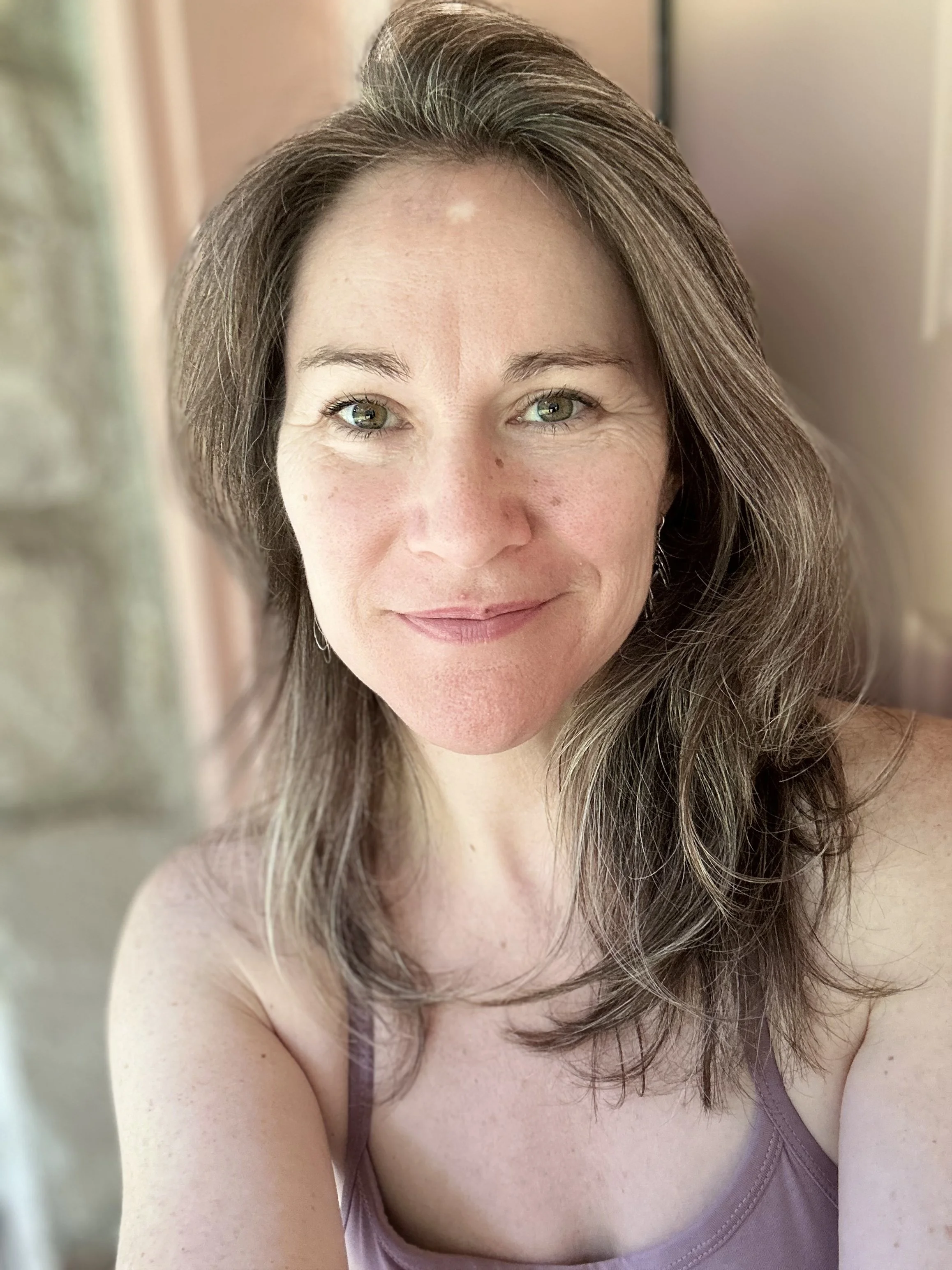 A woman with long brown hair and green eyes wearing a purple tank top, smiling, with blurred background.