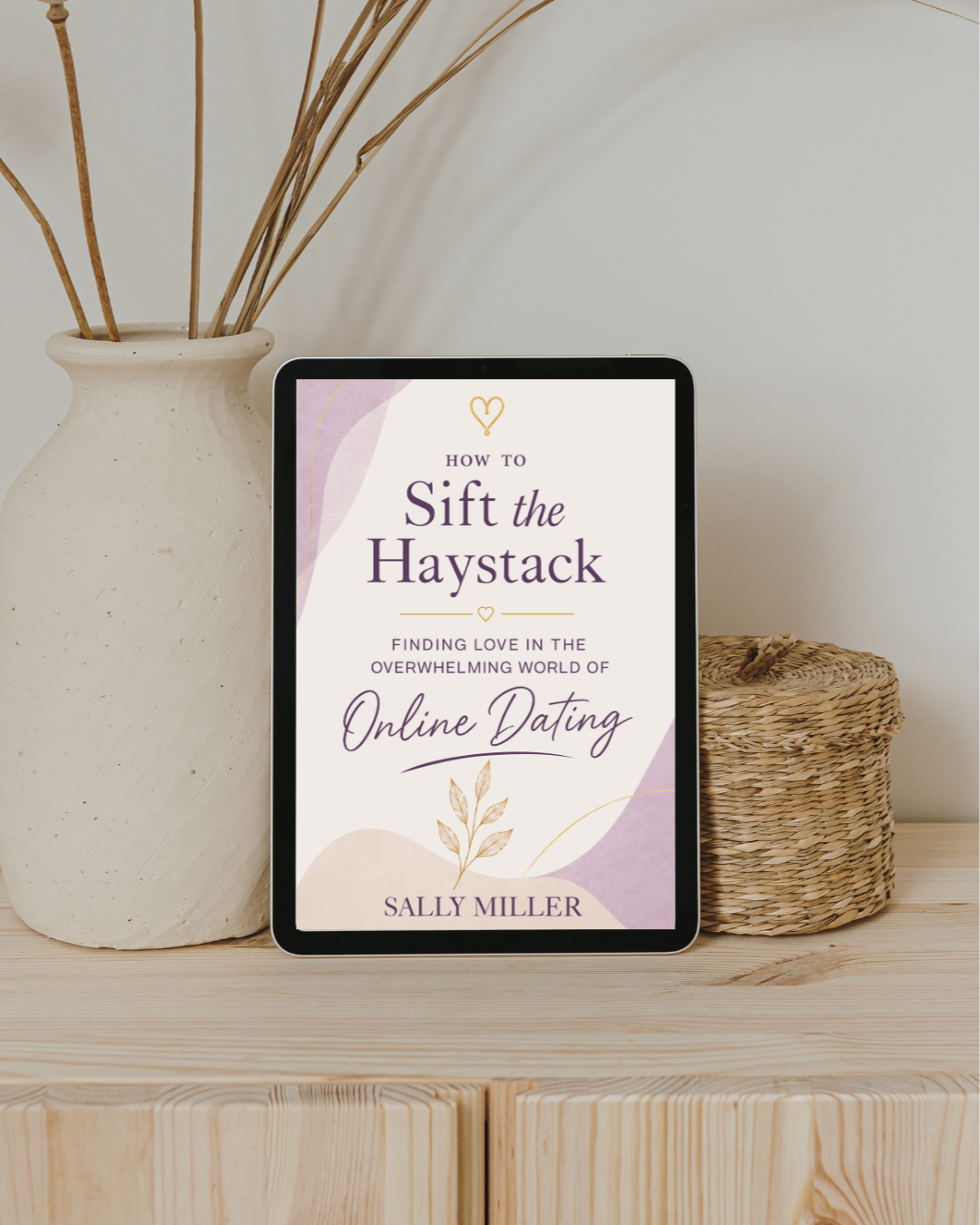Ebook titled 'How to Sift the Haystack: Finding Love in the Overwhelming World of Online Dating' by Sally Miller displayed on a tablet, placed on a wooden surface next to a large white ceramic vase with dried sticks and a woven basket.